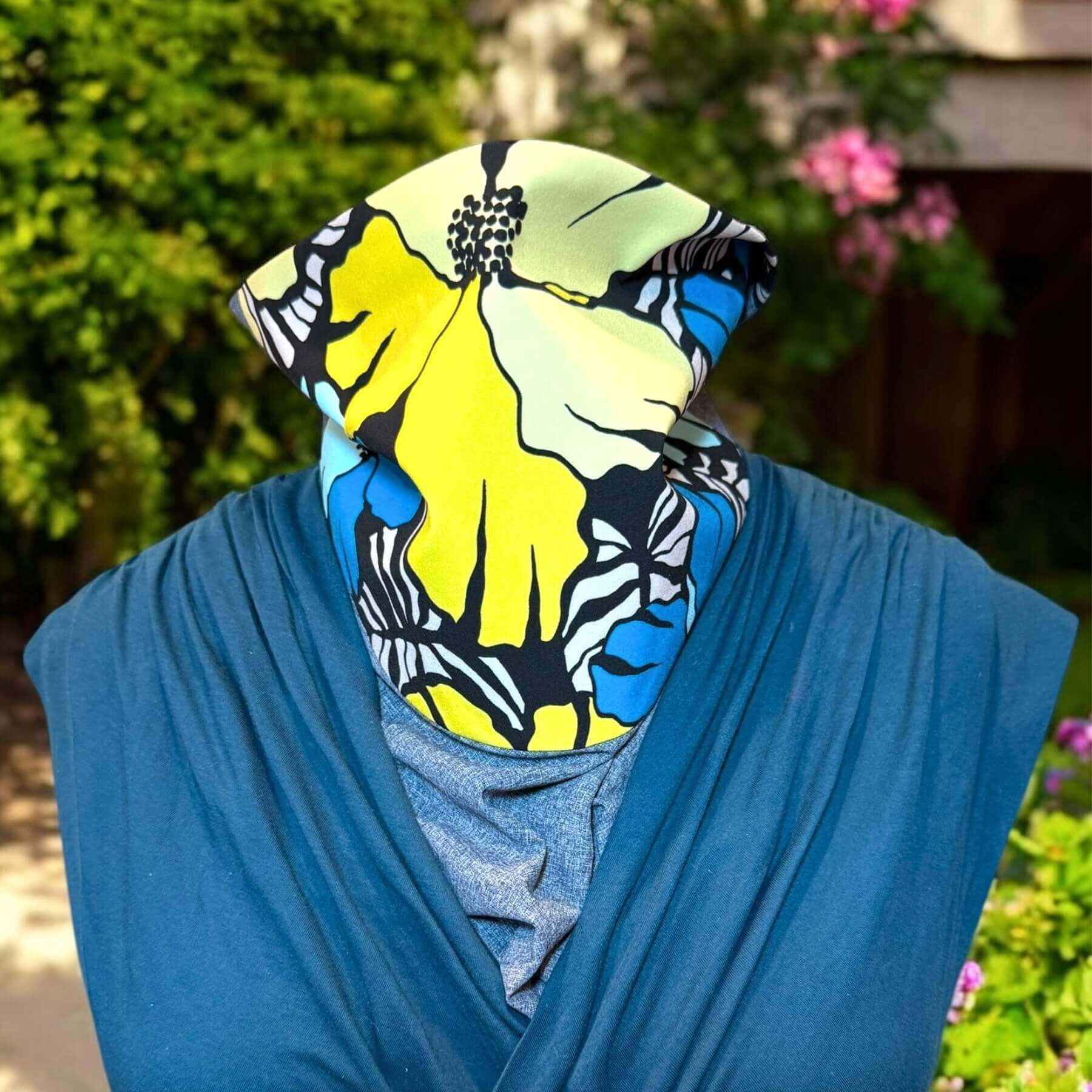 Stylish blue, yellow, black and white floral UV Sun Dickey paired with teal blue v-neck blouse. In background is backyard with trees and pink flowers