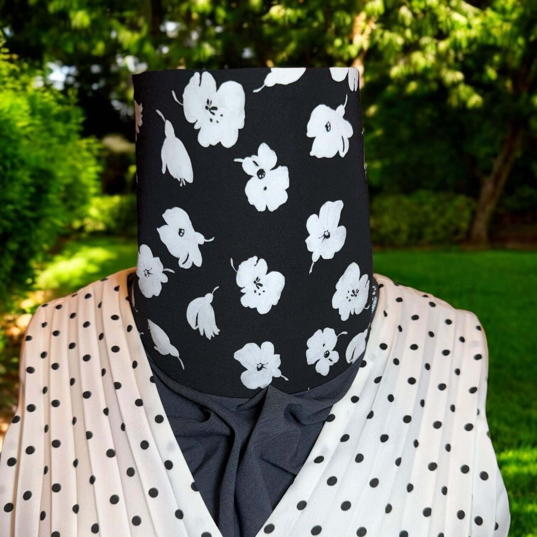 Stylish Sun Dickey for neck and chest UPF protection shown on mannequin in charcoal gray melange with white painted flowers on black is paired with black and white polka dot V-neck accordian pleated blouse. Behind in backyard is grass, trees and shrubs.