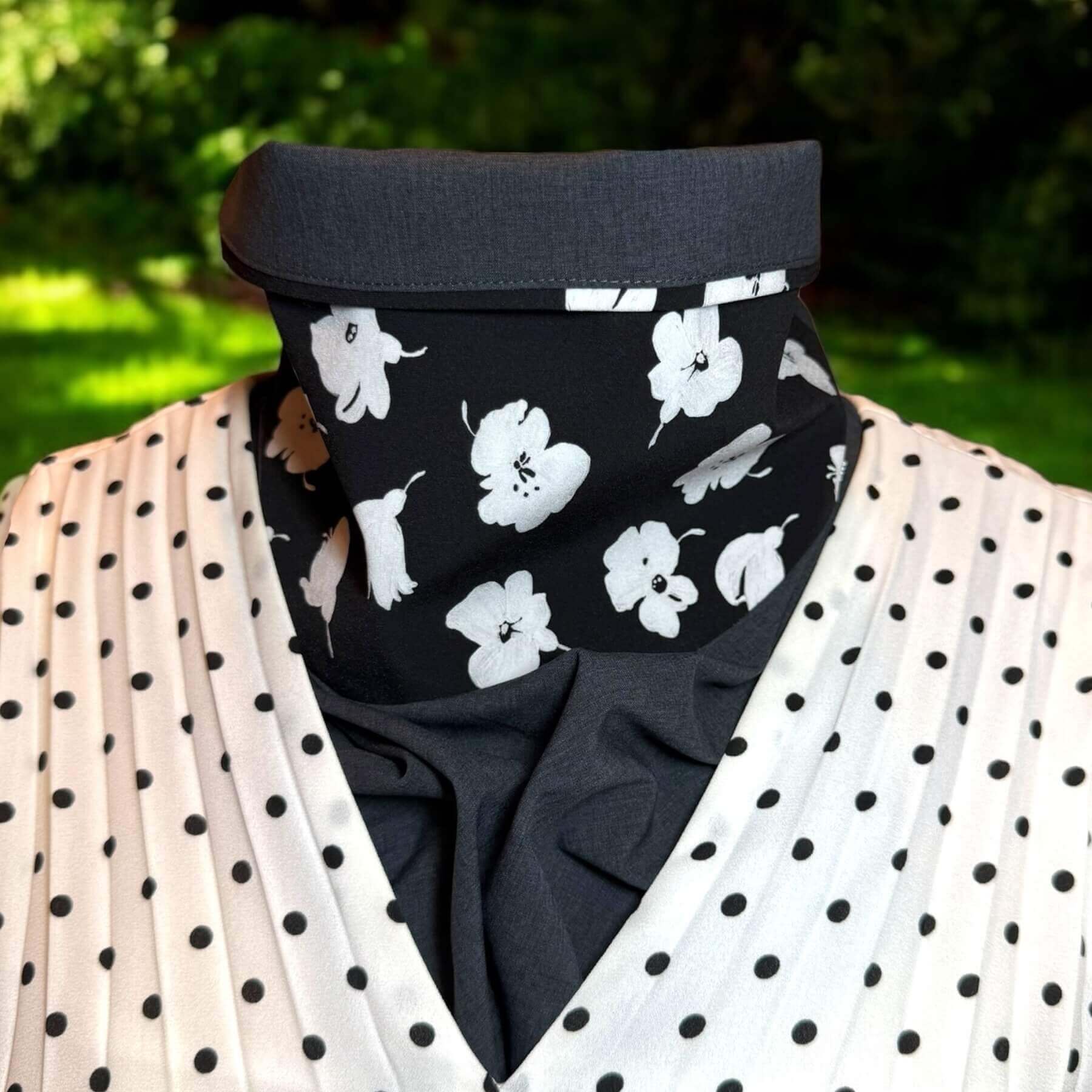 UPF protective Sun Dickey shown on mannequin with contrast collar folded down in charcoal gray melange with white painted flowers on black is paired with black and white polka dot V-neck accordian pleated blouse. Behind in backyard is grass with shrubs and a little sunlight.