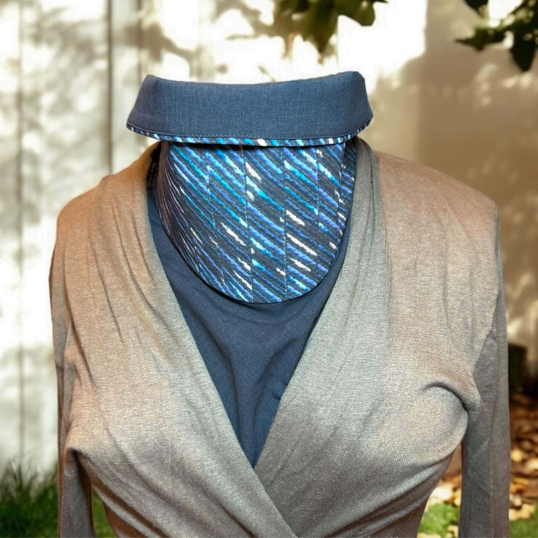 Sun Dickey with collar folded over shown on mannequin in charcoal gray melange with shades of blue, teal and white 'embroidered' (print) stripes in a diagonal pattern, paired with a beige sweater. In background is side of house in backyard with a few tree leaves and grass.