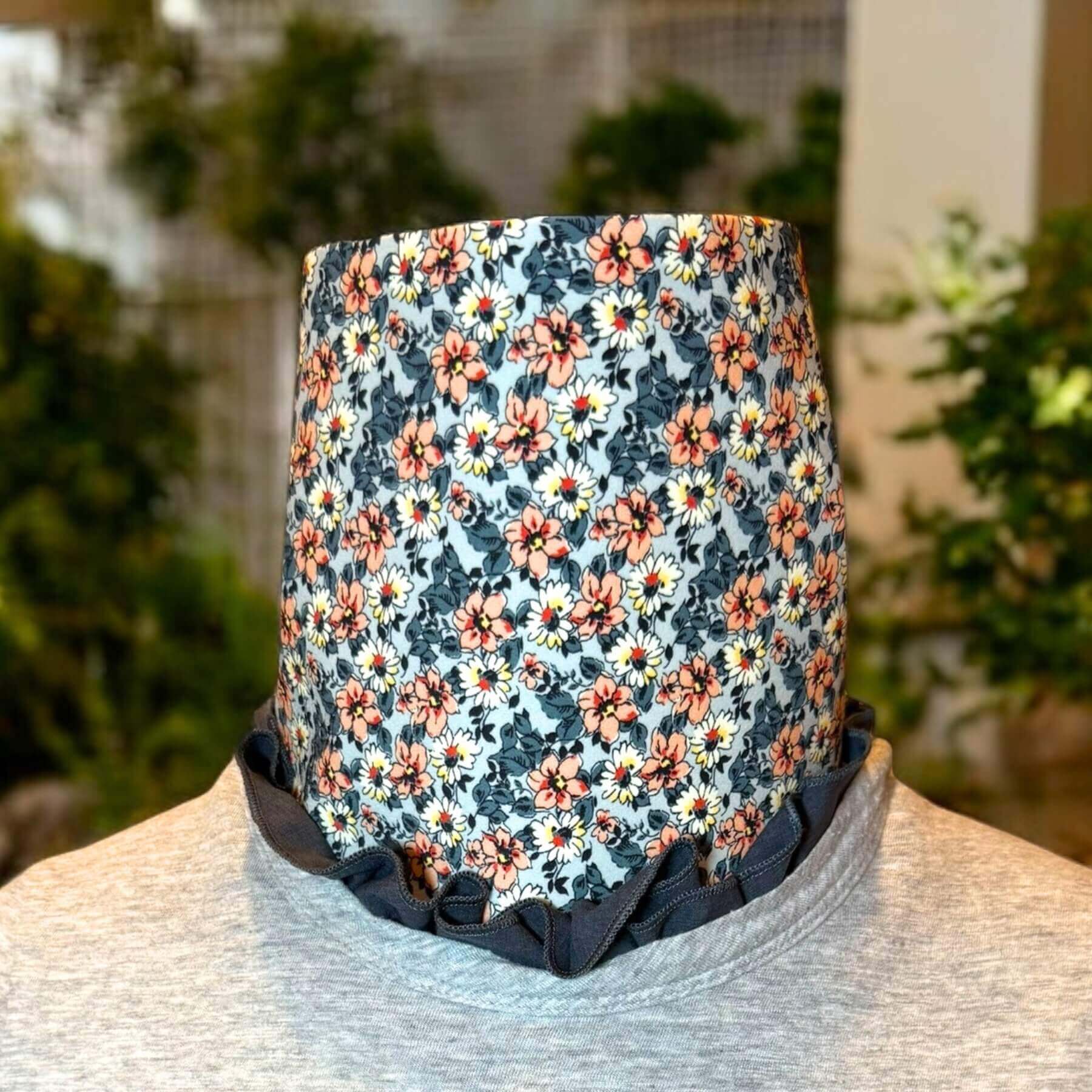 Fashion Sun Dickey shown in charcoal gray melange with a blue, peach and white floral print is shown with bib of dickey ruffled and peeking out of collar of gray t-shirt. In background is a house with green shrubbery