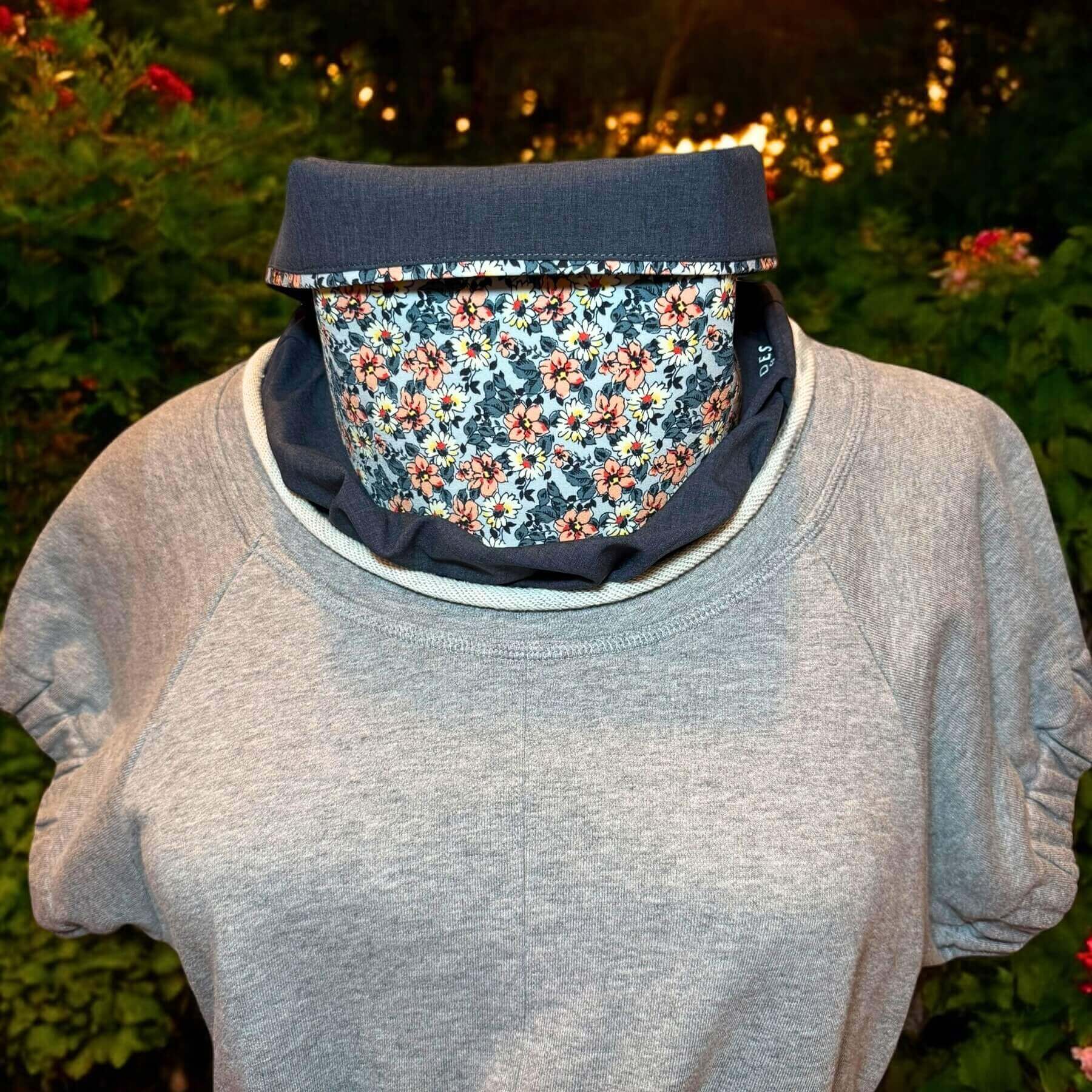 Stylish Sun Dickey shown in charcoal gray melange with a blue, peach and white floral print. The bib of the dickey is shown ruffled, peeking out of a Philip Lim gray top with a jewel rolled neckline. In background is foliage with red blooms and some sunlight.