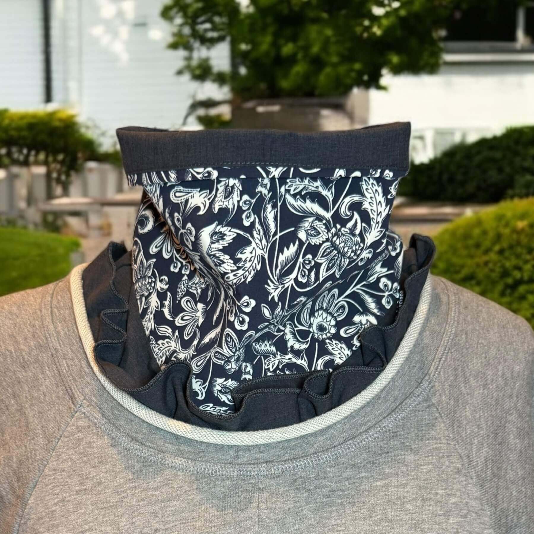 Stylish UV Sun Dickey shown on mannequin in blue melange with intricate navy blue and white stylized floral pattern with leaves, flowers and scrolls. The dickey bib is tucked into a gray Philip Lim sweatshirt and is seen ruffled, peeking out of the rolled jewel collar. Behind is house with backyard.