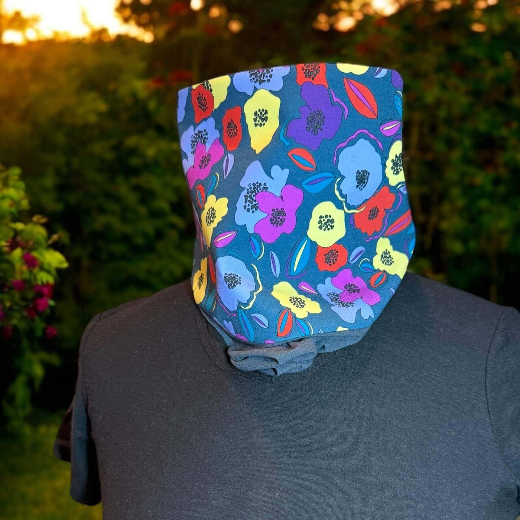 Chic UV Sun Dickey shown in charcoal gray melange with blue, violet, yellow and red floral print paired with black James Perse t-shirt. Behind in the backyard is green foilage and red flowers