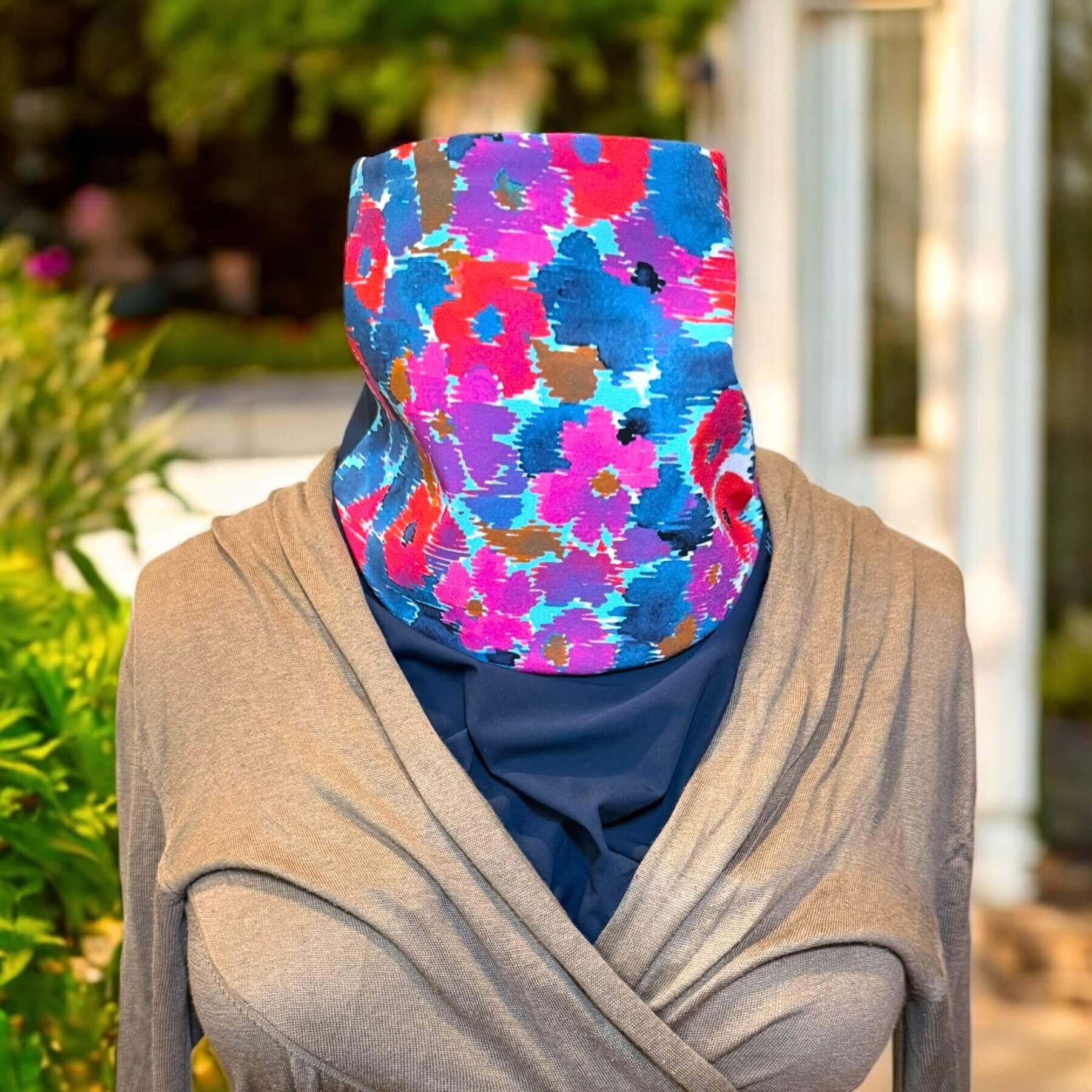 Chic Sun Dickey shown with collar up in navy blue with shades of blue, purple, violet and red floral print is styled with a lightweight beige wrap sweater. In the background is a white house, fence and backyard shrubs