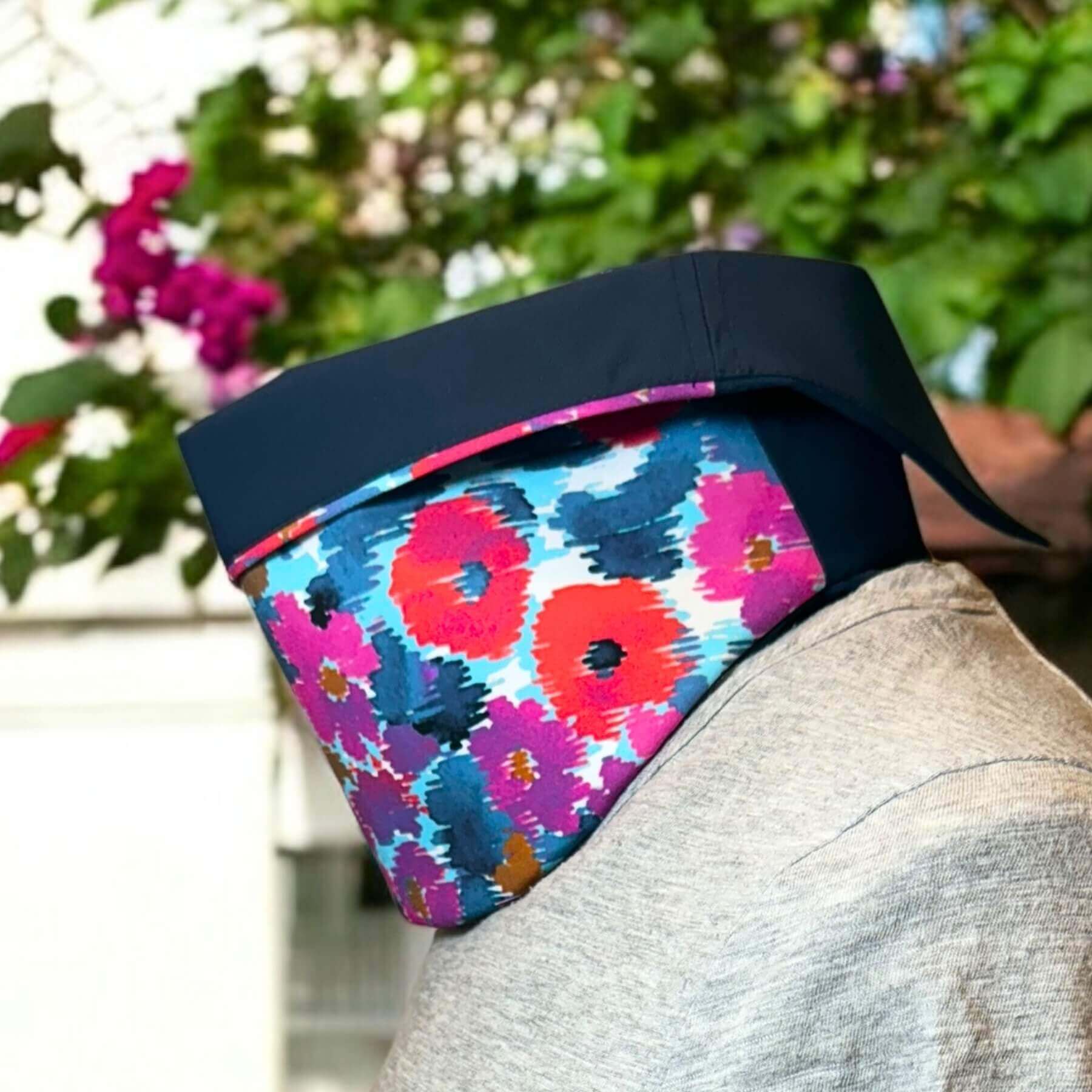 Side view of cute Sun Dickey shown with collar folded down in navy blue with shades of blue, purple, violet and red floral print paired with gray t-shirt. In the background is a white house and a tree limb with green leaves and pink flowers