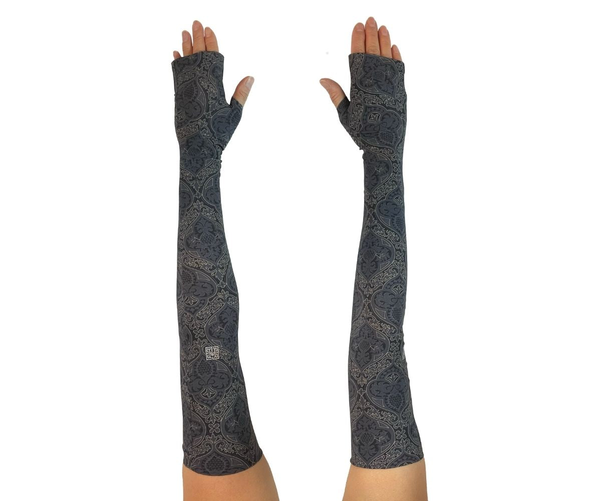 Heliades UPF 50 Sun Protective Clothing UV Arm Sleeves in fashionable, elegant allover black and gold print on a charcoal gray color. UV covered arms and hands shown outstretched.