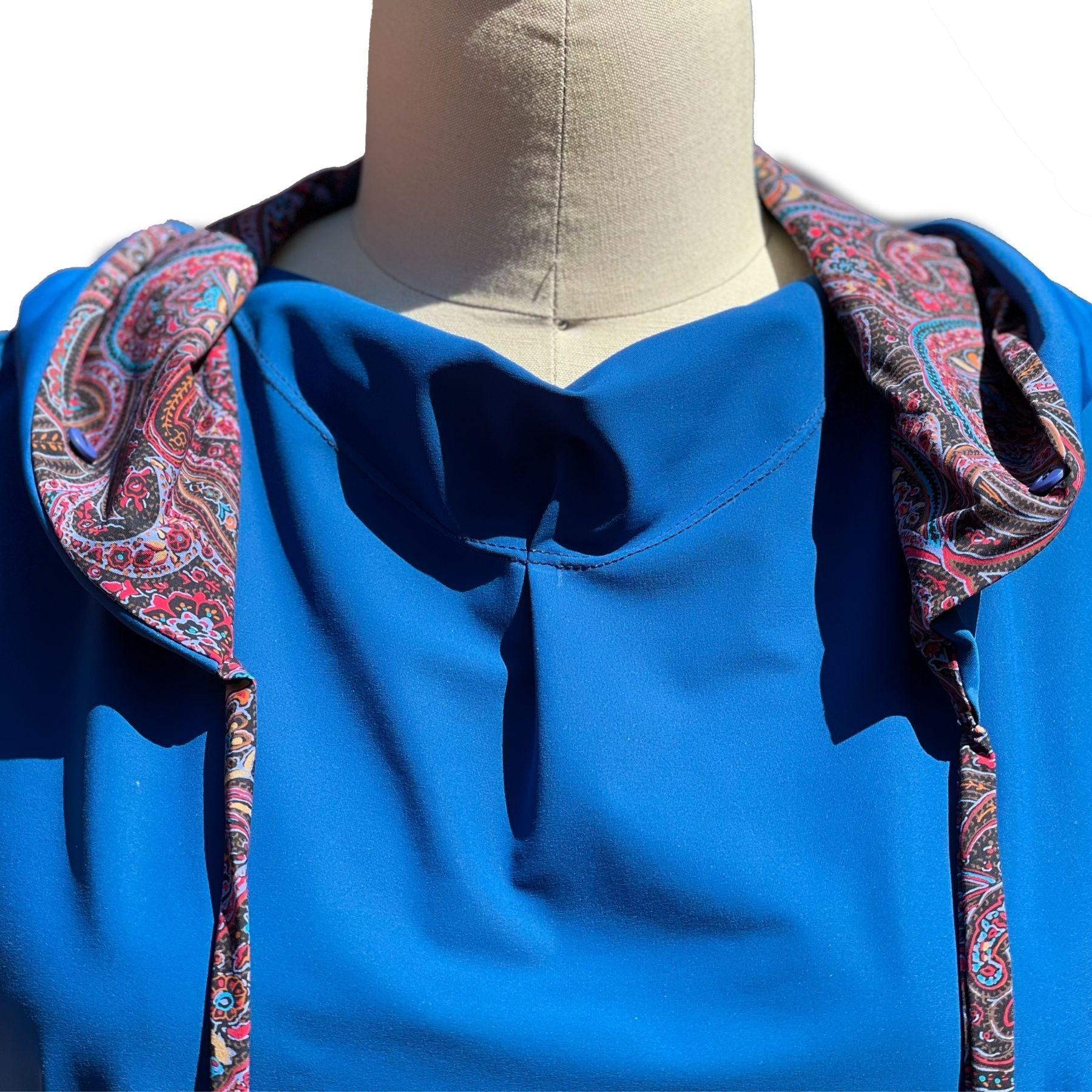 Close up view of UPF 50+ hoodie with collar draped in a jewel neckline. The hoodie is deep ocean peacock blue color and the sleeves & inside of the hoodie are all sewn in a paisley print with pink, red, lavender, turquoise colors.. Hidden, seamless button holes on the collar attach to the hoodie for even more neck coverage. The strings help secure the hood on windy days.