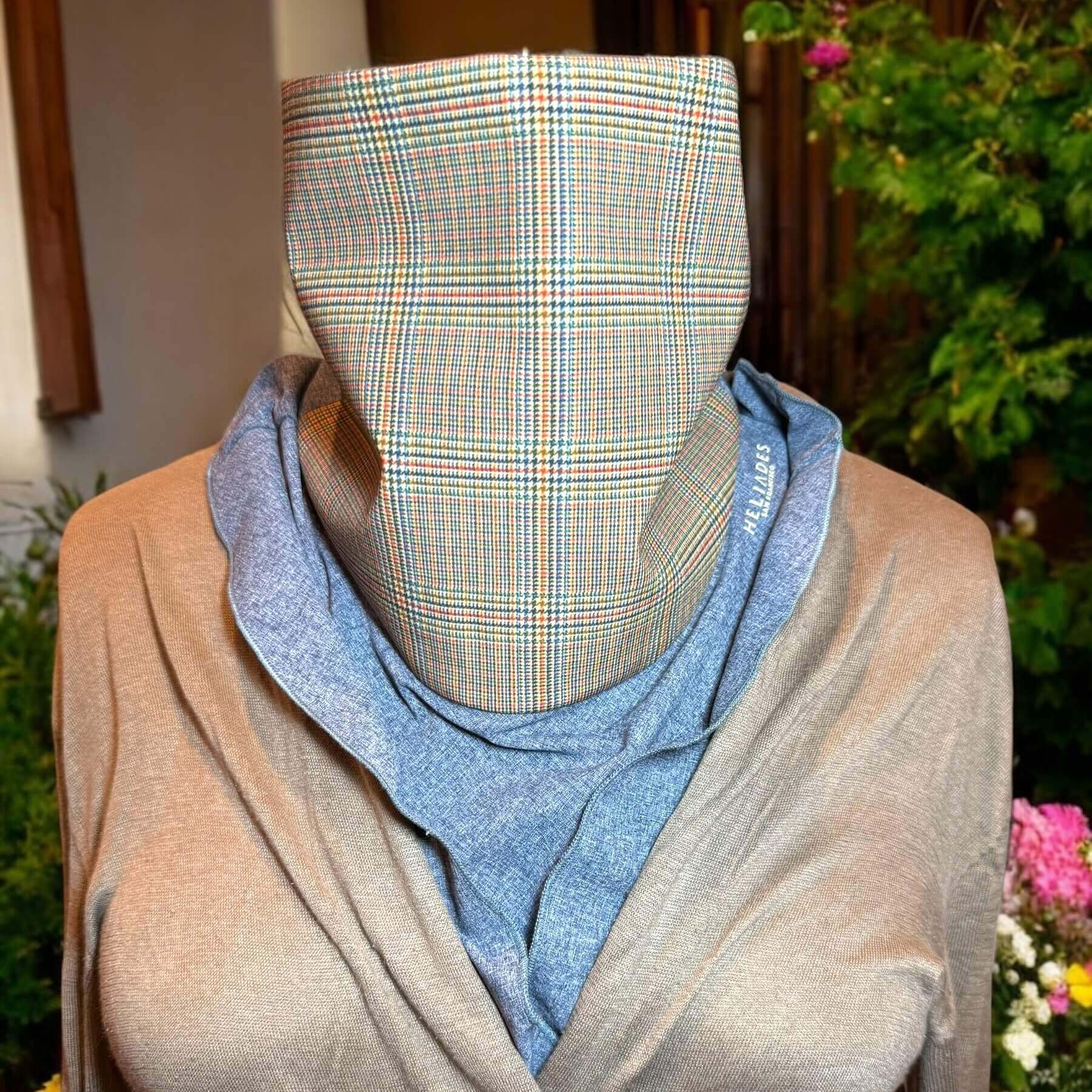 Pretty Sun Dickey shown on a mannequin in blue melange with classic glenn plaid print in shades of yellow, blue, orange and green is styled with a beige sweater. The bib of the dickey is ruffled, peeking out from the neckline of the sweater. In the background is a house with shrubs and flowers.