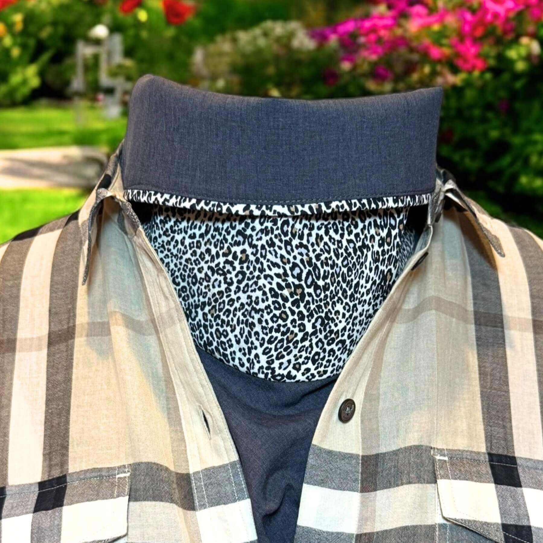 Sun Dickey with collar folded halfway down shown in charcoal gray melange with fabulous animal pattern in shades of cream, black, brown, is styled with classic Burberry plaid shirt in shades of beige, black and white. In background is the yard with grass, dirt, green bushes and red blooms
