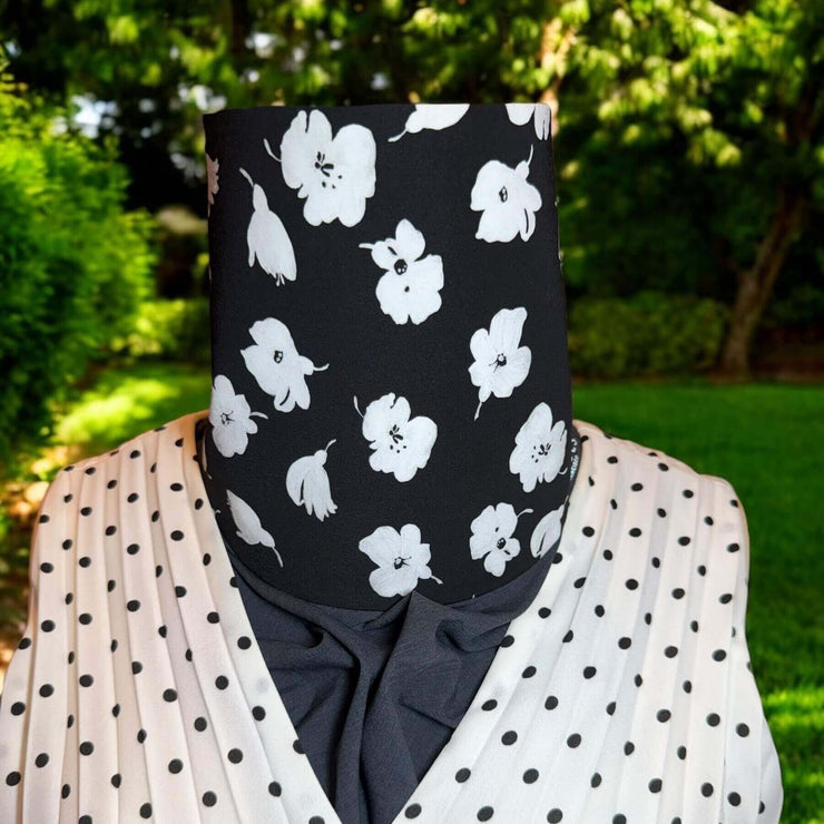 Stylish Sun Dickey for neck and chest UPF protection shown on mannequin in charcoal gray melange with white painted flowers on black is paired with black and white polka dot V-neck accordian pleated blouse. Behind in backyard is grass, trees and shrubs.