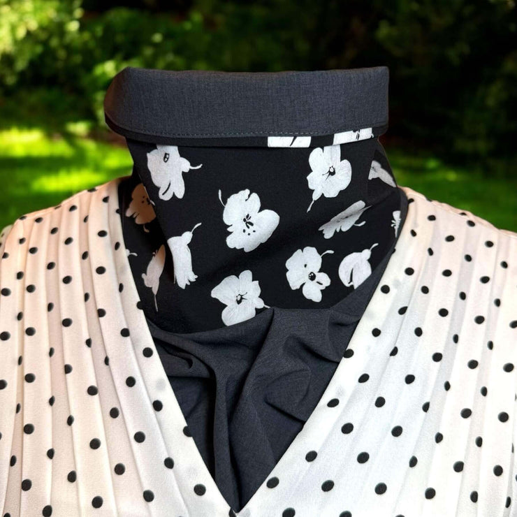 UPF protective Sun Dickey shown on mannequin with contrast collar folded down in charcoal gray melange with white painted flowers on black is paired with black and white polka dot V-neck accordian pleated blouse. Behind in backyard is grass with shrubs and a little sunlight.