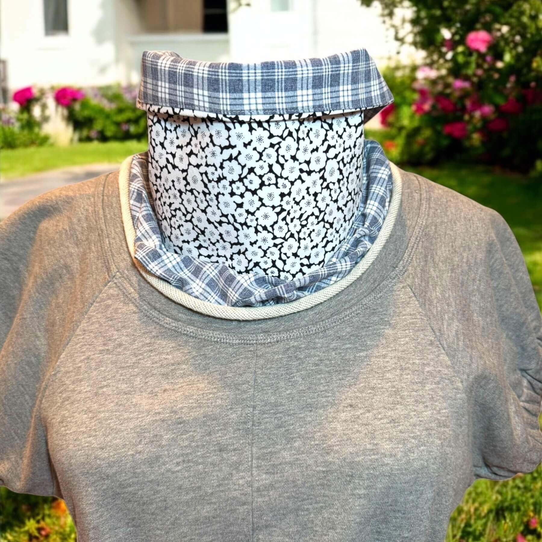 Fashion UV Sun Dickey shown in gray and white plaid with black, white floral print is tucked in to a Philip Lim short sleeve sweatshirt. The bib of the dickey is ruffled around the jewel neckline of the shirt. In background is a white house with grass and low shrubs and pink flowers.
