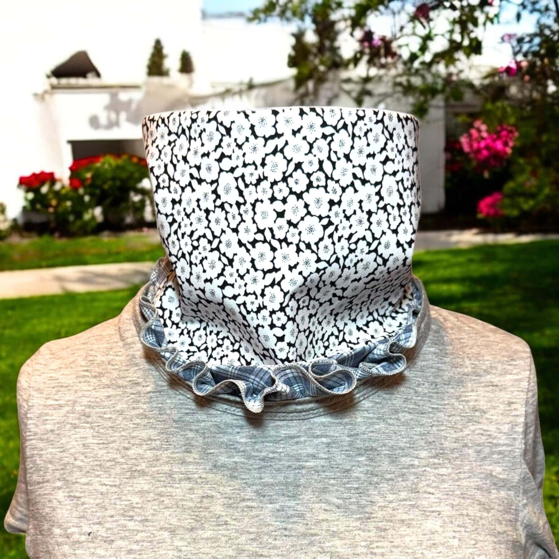 Stylish Sun protective Dickey shown in gray and white plaid with black, white floral print. The bib of the dickey is ruffled around the collar of a gray t-shirt. In background is a white house with grass , trees and low shrubs and pink flowers.