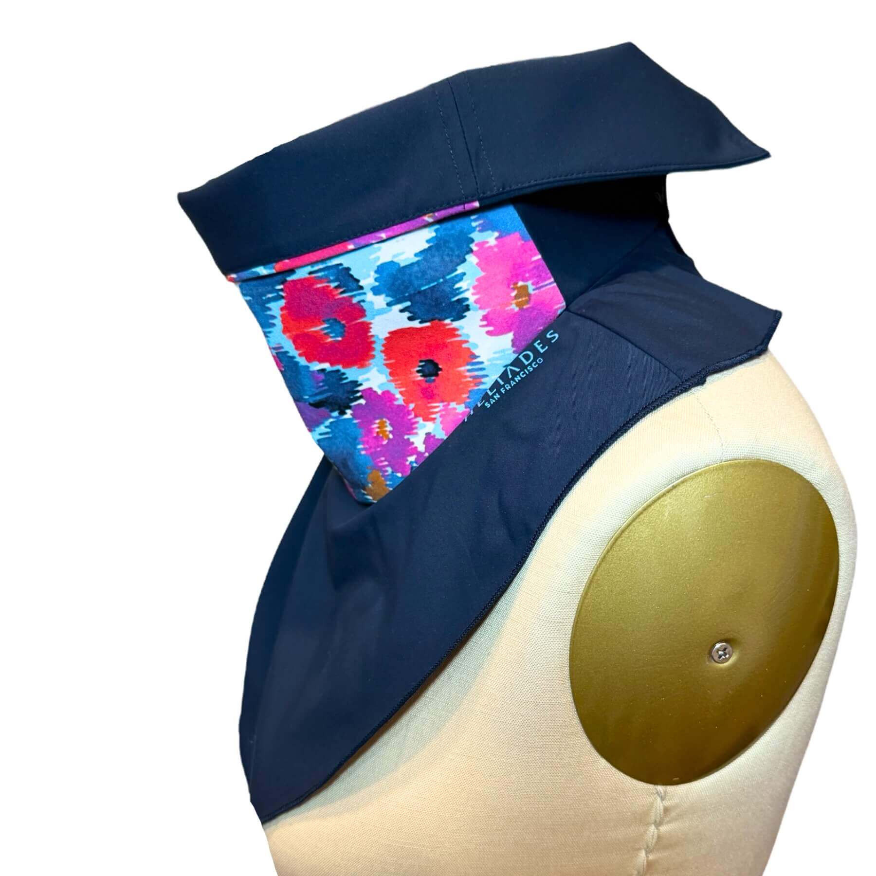 Side view of Sun Dickey shown with collar folded down in navy blue with shades of blue, purple, violet and red floral print. The reflective Heliades logo is glowing from flash photography