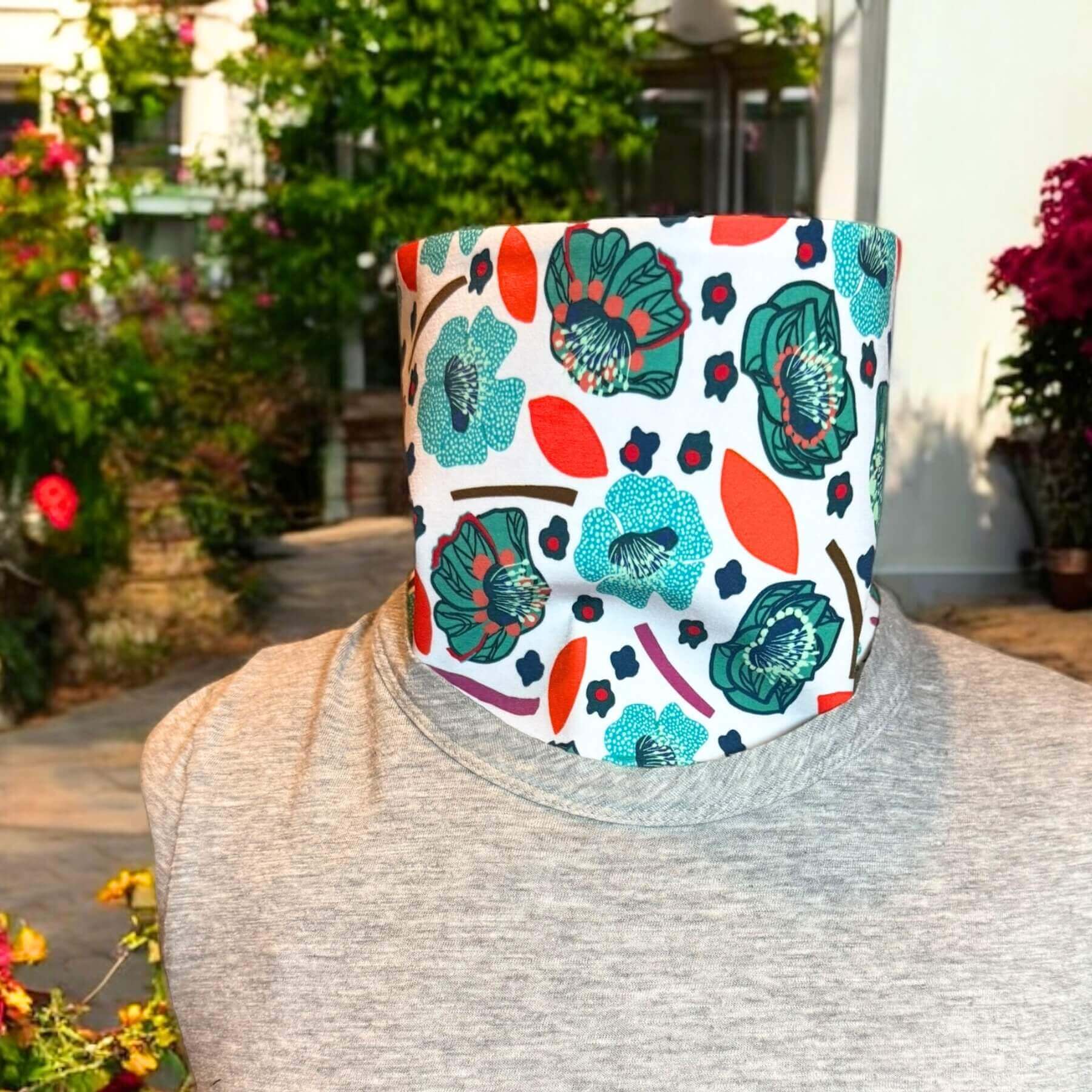 Sun Dickey in navy blue with detailed teal, blue, mint and red flower motif on white print shown with bib tucked into a gray t-shirt. Behind is a white house with green foliage, concrete path and red flowers and leaves