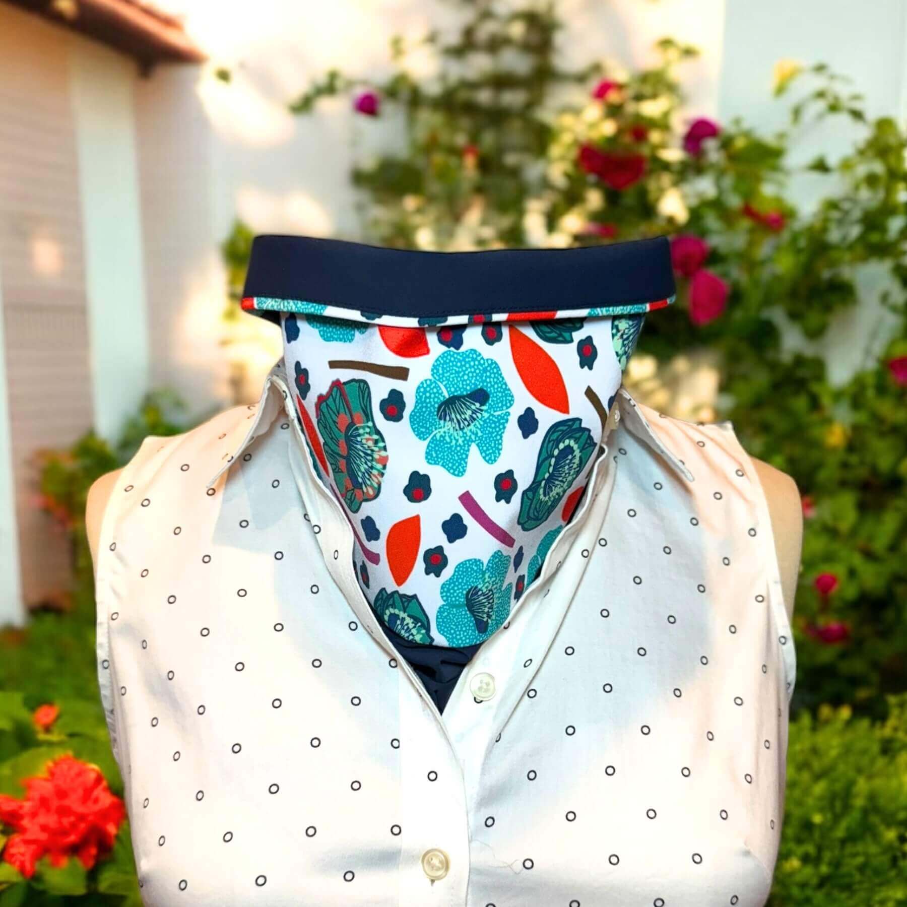 Sun Dickey in navy blue with detailed teal, blue, mint and red flower motif on white print is shown with collar turned down one inch to reveal navy blue lining contrast, is paired with a black and white polka dot collared dress shirt. Behind in background is green foliage trailing up a white garage wall. There are orange, yellow and red blossoms and grass.