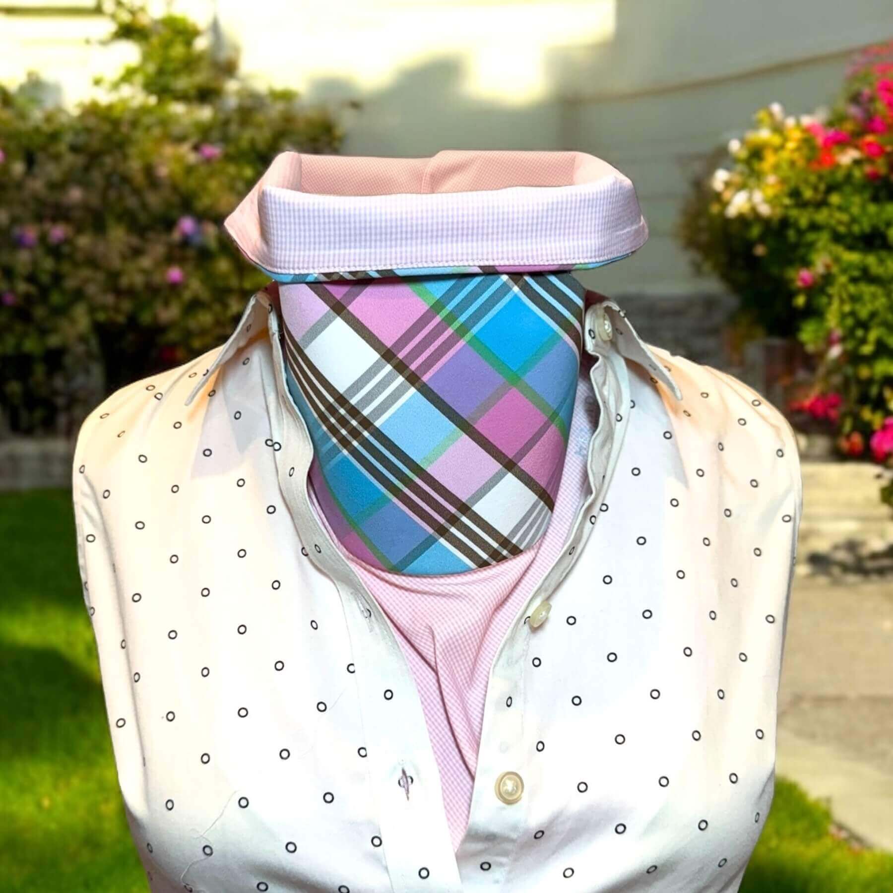 Sun Dickey shown with top of collar turned down in pink, white micro check with pink, blue, green and white plaid, styled with white and black polka dot collared dress shirt. In background is white house, pink, red and white blooms on shrubs, grass.