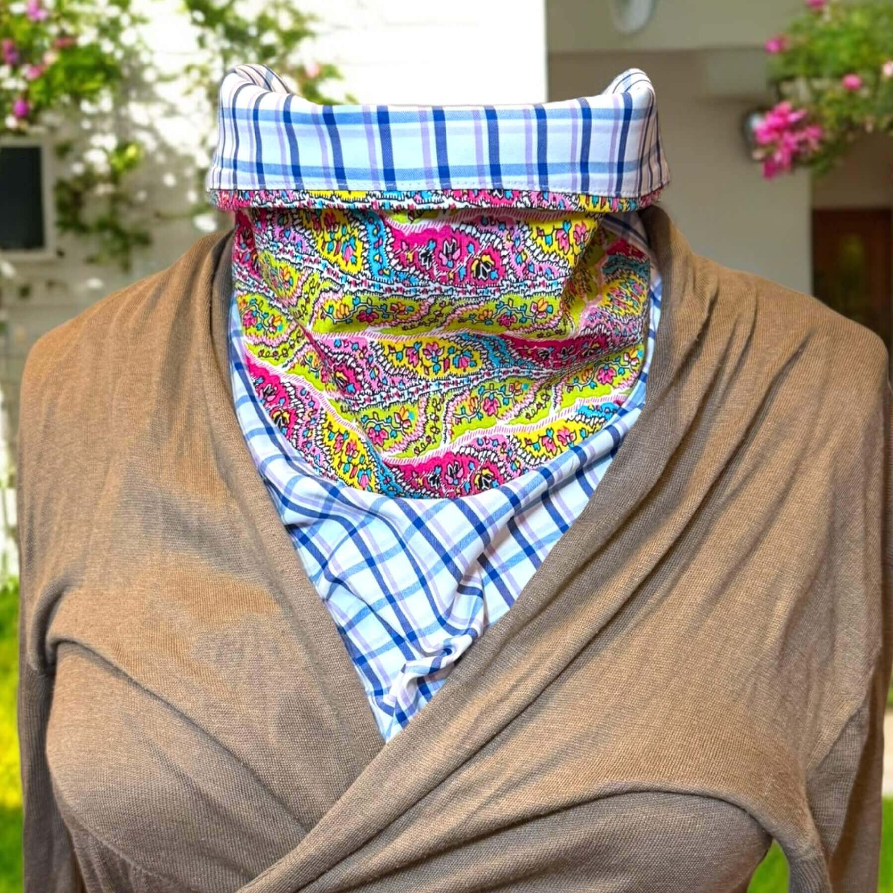 Sun Dickey in blue, white plaid with pink, blue, green paisley print shown with collar folded and styled with lightweight, beige wraparound sweater. In background is white house with green shrubs, pink flowers.