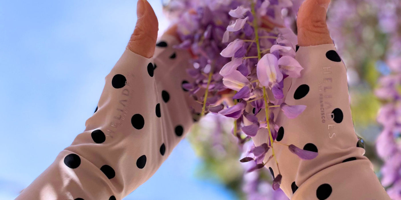 Woman wearing Heliades pink polka dot sun protection gloves, holding wisteria, showcasing fashionable UV protective clothing.