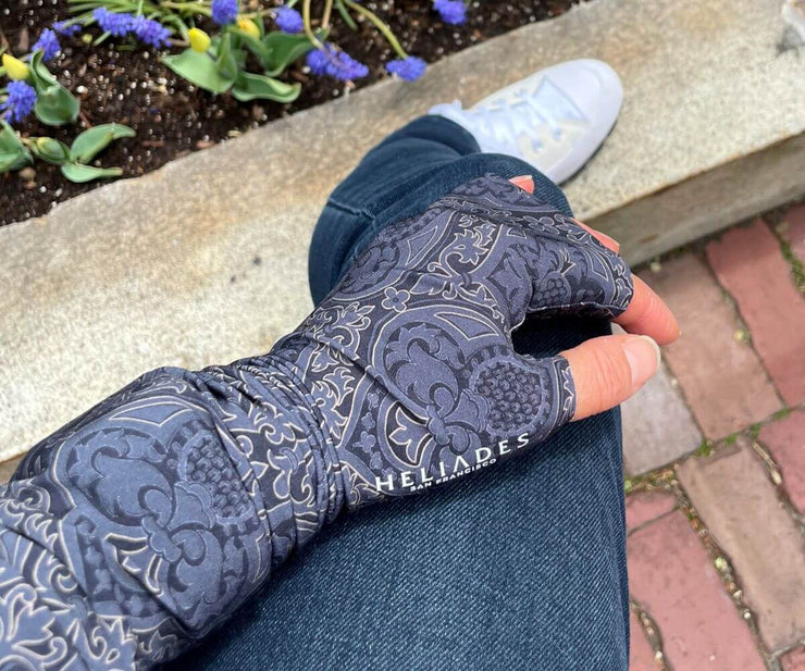 Heliades UPF 50 Sun Protective Clothing UV Arm Sleeves in fashionable, elegant allover black and gold print on a charcoal gray color. UV covered arm and hand rests on jeans, showing silver reflective logo trim with iWatch and bracelet fits underneath sleeves.