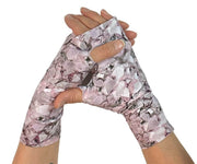 Heliades UPF 50 Sun Protective Clothing UV sun gloves on printed fabric. Shown is fashionable, elegant allover pink, lavender, white, magenta print. UV sun covered hands shown clasped together with metallic pink logo trim