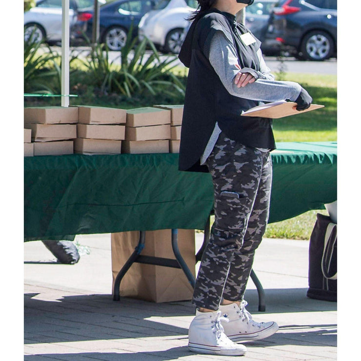 Woman wearing UPF 50+ Hoodie as pullover tunic over a gray shirt and camo pants. The tunic is longer in back for extra rear coverage. She is wearing white Converse high tops, UPF 50+ black gloves and holding a clipboard. Photo credit: Karen Ambrose Hickey