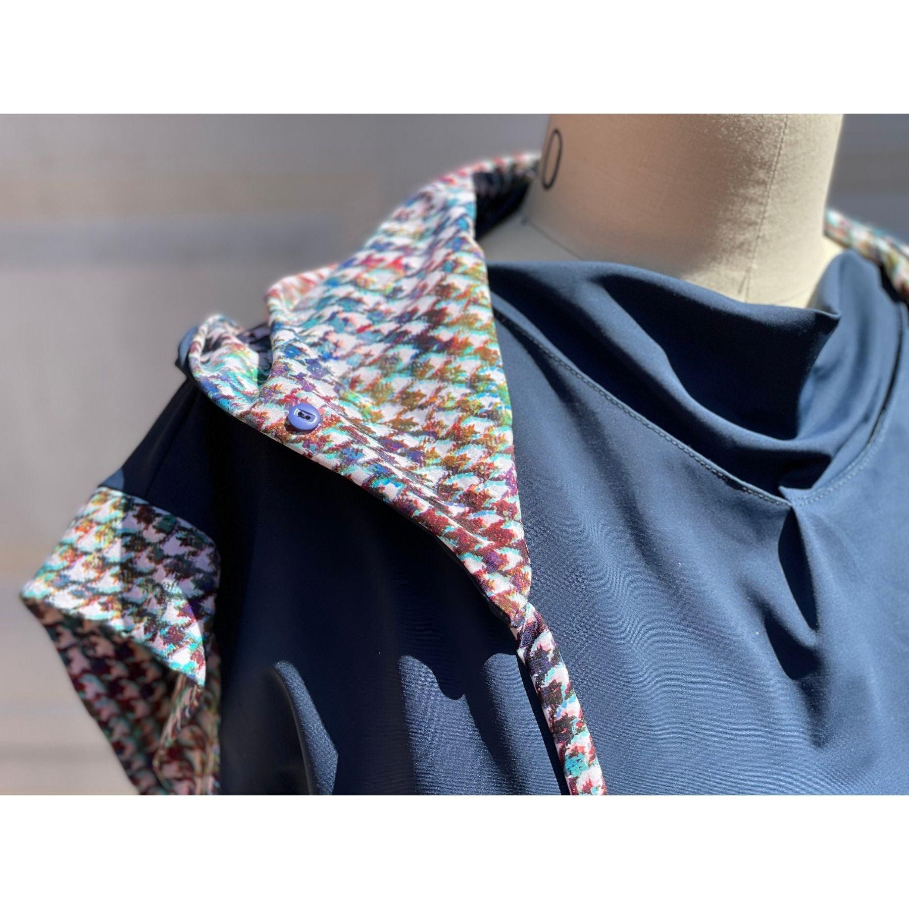 Close up view of UPF 50+ hoodie with collar unfolded and extended up to create extra neck coverage from the sun’s UV rays. The hoodie is true navy blue and inside of the hoodie and string ties are all sewn in painterly houndstooth plaid print. Hidden, seamless button holes on the collar attach to the hoodie for even more neck coverage. The strings help secure the hood on windy days.