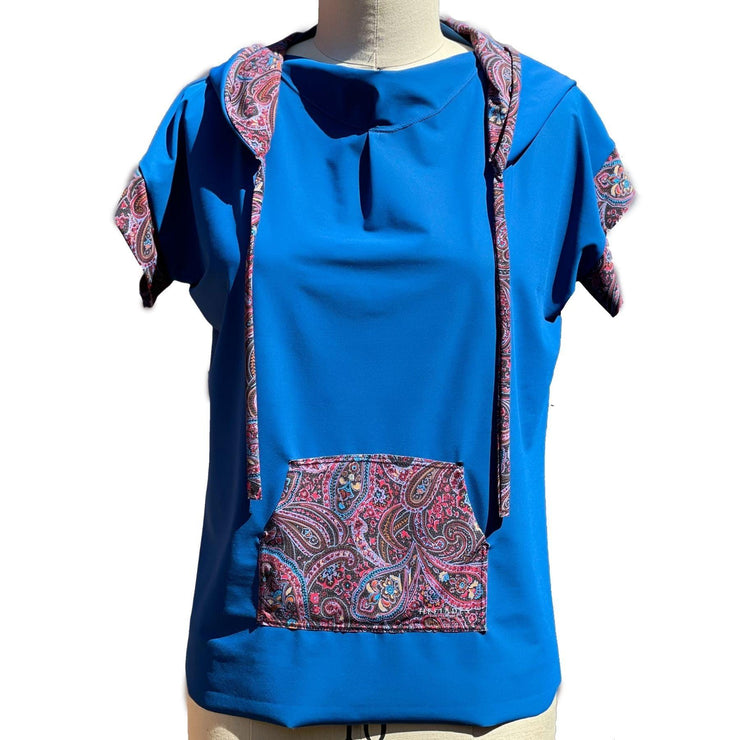 A short sleeved, UPF 50+ hoodie tunic is placed on a mannequin. The hoodie is deep ocean peacock blue color and the sleeves & inside of the hoodie are all sewn in a paisley print with pink, red, lavender, turquoise colors. The collar is folded down and draped, making nice folds around the jewel neckline. The Hoodie is long, loose and flowing like a tunic with split vents at the hips