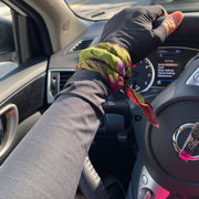 An arm on steering wheel wearing a UPF 50 sun protective UV arm sleeves in black with pink metallic logo and a bright green multi color scarf wrapped around wrist