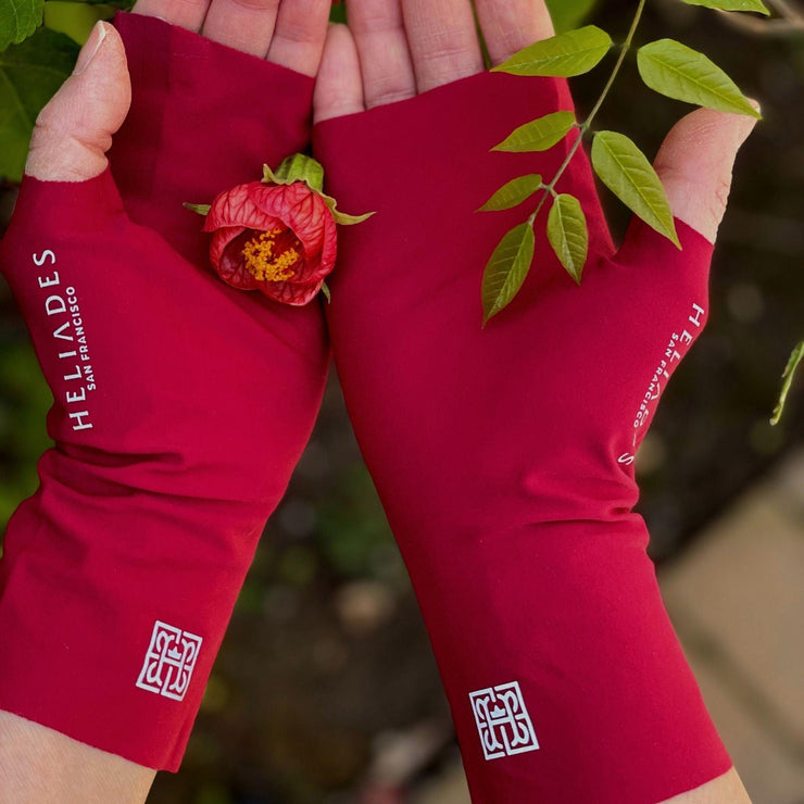 Hands cradle a little flower with salmon and red petals and orange center. Leaves in back ground. On hands are pair of UPF 50+ fingerless sun protection gloves in deep red with silver reflective logos