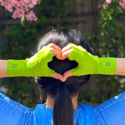 Woman with black haired ponytail and blue shirt holds hands behind her head in a heart shape. On hands is pair of UPF 50+ fingerless sun protection gloves in bright, grassy green color with black logos. In background are pink flowers